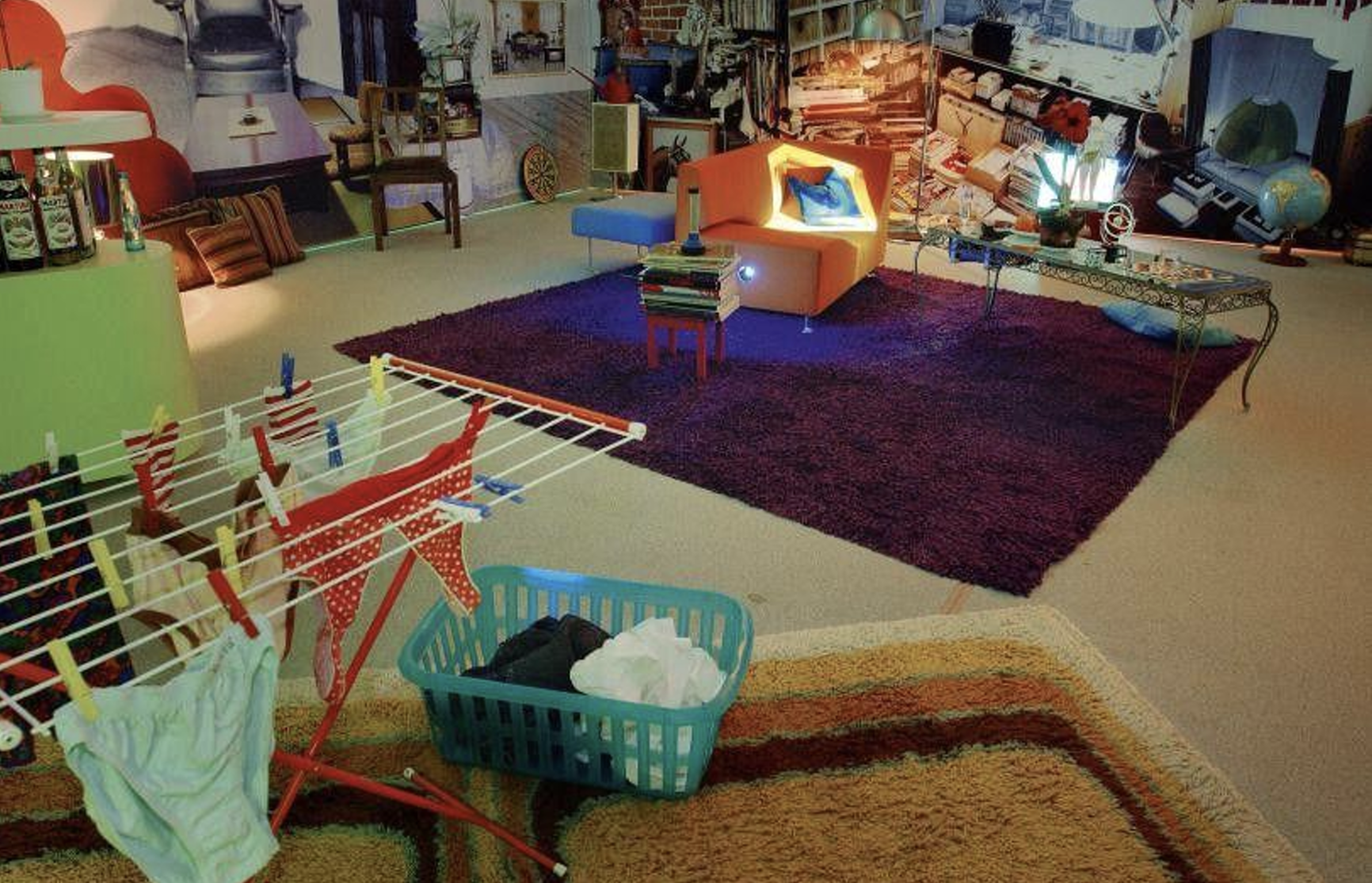 Buntes Wohnzimmer mit lila Teppich, orange Sitzgelegenheit, Wäscheständer und Wäschekorb